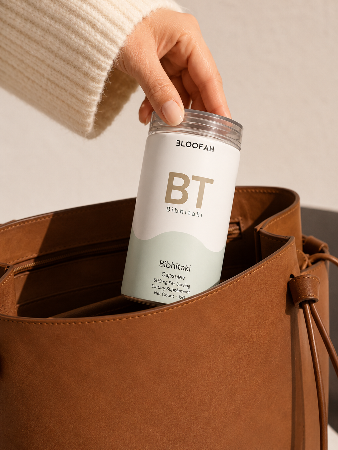 A hand wearing a cream knit sleeve holds a clear cylindrical bottle labeled BLOOFAH BT Bibhitaki capsules, placing it into a brown suede bucket bag.